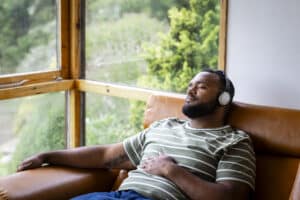 A mid adult man listening to music through wireless headphones and sitting back on a leather sofa in his living room at home in Hexham, North East England. He is listening to the music while looking content with his eyes closed.